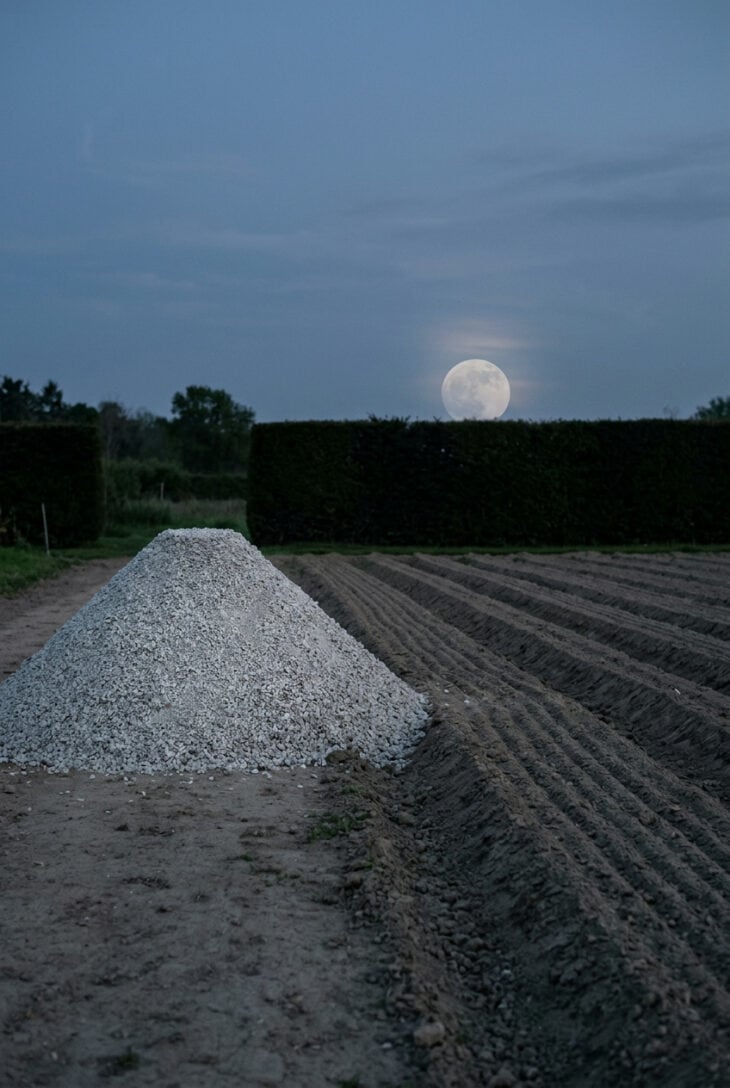 Flat-topped cone of pale gravel beside raked gravel at dusk with a full moon rising behind a dark hedge